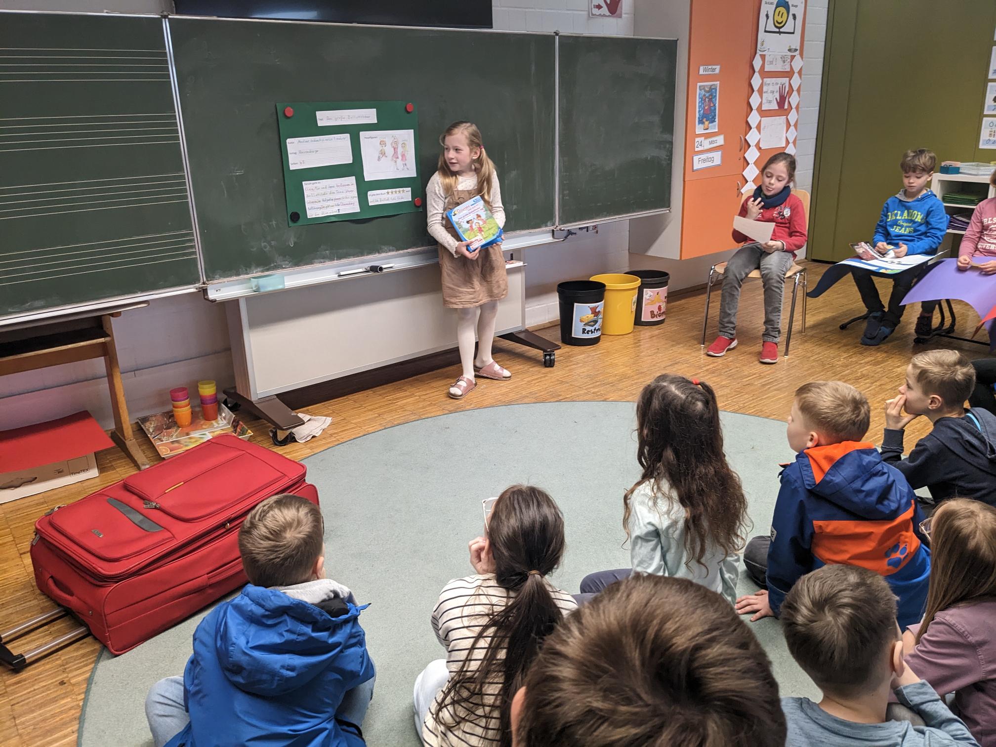 Ein Kind stellt ein Buch an der Tafel vor. Die Klasse beobachtet sie aufmerksam.